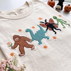 May include: A cream-coloured t-shirt featuring embroidered designs of a brown Bigfoot holding a pink flower, a teal dinosaur, a black moth with red eyes, and a green alien, all surrounded by small orange star shapes. The shirt has a Comfort Colors label.