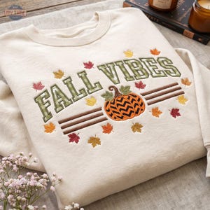 Op de afbeelding: Een crèmekleurige sweater met ronde hals en geborduurde tekst FALL VIBES in een groen ruitpatroon. Onder de tekst staat een oranje pompoen met een zwart chevronpatroon, omringd door kleine geborduurde herfstbladeren in rood, oranje en geel.
