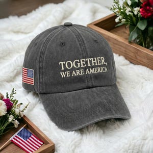 Może przedstawiać: Czapka bejsbolowa w kolorze ciemnoszarym z białym napisem "TOGETHER, WE ARE AMERICA". Z boku naszywka z flagą amerykańską. Czapka leży na białej, puszystej powierzchni.