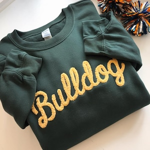 May include: A dark green sweatshirt with the word "Bulldog" in a gold, textured font across the chest. The sweatshirt has a crew neck and long sleeves with ribbed cuffs. Pom-poms are visible in the background.