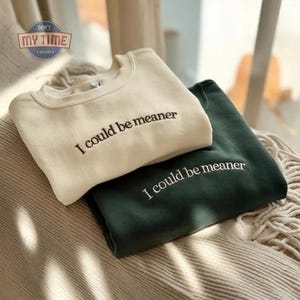 May include: Two crewneck sweatshirts, one cream-colored and one dark green, are stacked on top of each other. The phrase "I could be meaner" is embroidered in black on the front of each sweatshirt. The sweatshirts are resting on a textured, neutral-toned surface.