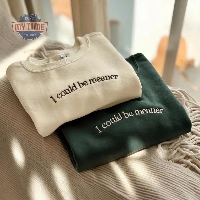 May include: Two crewneck sweatshirts, one cream-colored and one dark green, are stacked on top of each other. The phrase "I could be meaner" is embroidered in black on the front of each sweatshirt. The sweatshirts are resting on a textured, neutral-toned surface.