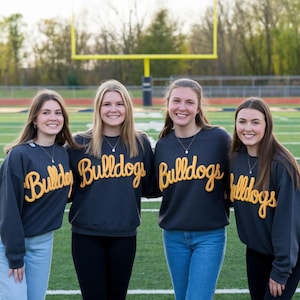 May include: Four people wearing dark gray sweatshirts with the word "Bulldogs" in yellow script. They are standing on a green field in front of a yellow goalpost. The sweatshirts are paired with blue jeans and black pants.