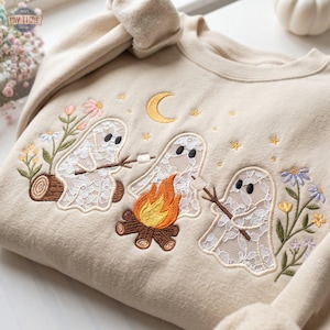 Könnte beinhalten: Ein beigefarbenes Sweatshirt mit Rundhalsausschnitt und einer Stickerei von drei weißen, spitzenartigen Geistern, die um ein Lagerfeuer sitzen. Die Geister rösten Marshmallows. Das Motiv zeigt einen gelben Halbmond, Sterne und verschiedene Blumen.