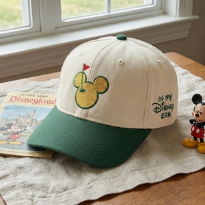 May include: Cream-coloured baseball cap with a green brim and a green button on top. The cap features an embroidered Mickey Mouse golf design and the text "IN MY Disney ERA" in black. A vintage Disneyland guide is visible.