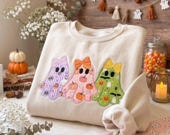 May include: A cream-coloured crewneck sweatshirt featuring a graphic of three ghosts wearing bows. The left ghost is purple with stripes, the middle ghost is pink with pumpkins, and the right ghost is green with bats and pumpkins.