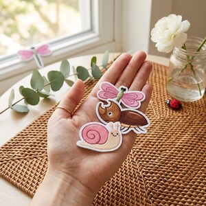 May include: Three embroidered patches featuring a pink dragonfly, a brown rhinoceros beetle, and a light beige snail with a pink shell. Each patch has a white border and detailed stitching, showcasing insect and mollusk designs in vibrant colors.