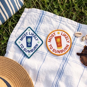 May include: Two embroidered patches featuring the text MOST LIKELY TO SUNBURN. One is a diamond shape with a blue border and a blue sunscreen bottle icon. The other is a circular shape with an orange border and an orange sunscreen bottle icon.