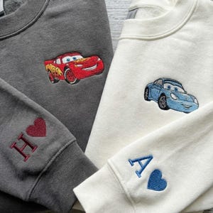 Könnte beinhalten: Zwei Sweatshirts, eines grau und eines cremefarben, jeweils mit einem Cartoon-Auto bestickt. Das graue Sweatshirt zeigt ein rotes Auto mit der Nummer 95. Das cremefarbene Sweatshirt hat ein blaues Auto. Ärmel haben Initialen und Herzen.