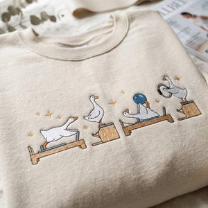 May include: A beige crewneck sweatshirt featuring an embroidered design of four white geese performing various Pilates exercises on wooden equipment, accented with small gold stars scattered around the figures.