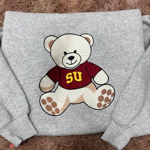 Op de afbeelding: Een grijze sweatshirt met een geborduurde teddybeer die een bordeauxrood shirt draagt met "SU" in geel. De beer heeft cr&egrave;mekleurige vacht en bruine accenten op zijn poten. De sweatshirt heeft lange mouwen.