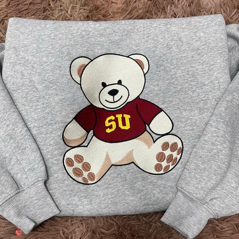 Op de afbeelding: Een grijze sweatshirt met een geborduurde teddybeer die een bordeauxrood shirt draagt met "SU" in geel. De beer heeft cr&egrave;mekleurige vacht en bruine accenten op zijn poten. De sweatshirt heeft lange mouwen.