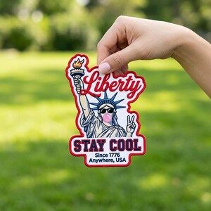 Pode incluir: Um emblema bordado recortado apresentando a Estátua da Liberdade usando óculos de sol e soprando uma bolha rosa. O texto diz Liberty, STAY COOL e Since 1776 Anywhere, USA. O emblema tem uma borda vermelha e um fundo branco.