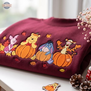 May include: A maroon crewneck sweatshirt featuring embroidered characters from Winnie the Pooh, including Piglet, Pooh, Eeyore, and Tigger, each sitting inside or next to orange pumpkins, surrounded by small embroidered autumn leaves.