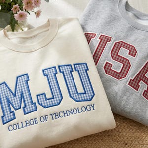 Può includere: Due felpe girocollo. Una è color crema con lettere a quadretti blu che compongono "MJU" e le parole "COLLEGE OF TECHNOLOGY" ricamate sotto. L'altra è grigia con lettere a quadretti rosse che compongono "TSA".