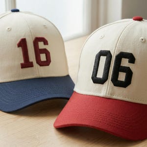 Op de afbeelding: Twee baseballpetten. Eén is crèmekleurig met een marineblauwe klep en het nummer 16 in bordeauxrood. De andere is crèmekleurig met een rode klep en het nummer 06 in zwart. Beide hebben een knoop bovenop.
