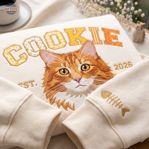 May include: A cream-coloured sweatshirt featuring an embroidered orange tabby cat face in the centre. Above the cat, the word COOKIE is stitched in yellow and orange patterned letters. The text EST. 2026 is present, and a fish skeleton is on the sleeve.