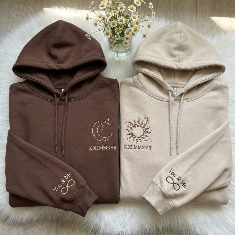 May include: Two matching hoodies, one brown and one beige, with embroidered celestial designs. The brown hoodie features a moon and stars, while the beige one has a sun. Both have the text "You & Me" and an infinity symbol on the sleeve.