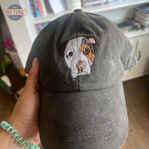 May include: A dark gray baseball cap with an embroidered image of a white and brown dog with a pink nose. The dog's eyes are brown and black.