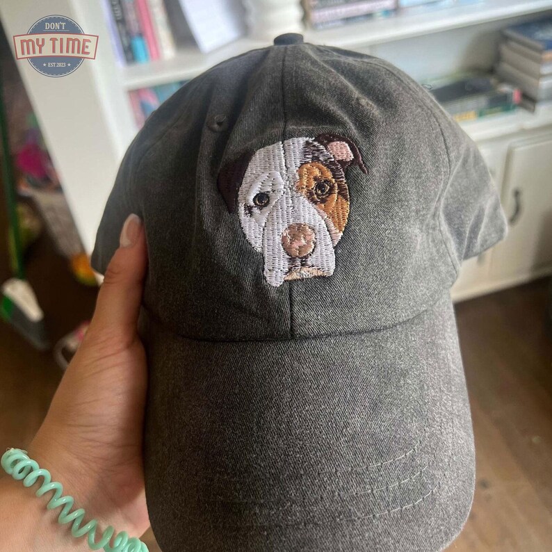 May include: A dark gray baseball cap with an embroidered image of a white and brown dog with a pink nose. The dog's eyes are brown and black.
