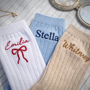 Op de afbeelding: Drie geribbelde sokken in wit, lichtblauw en beige. De witte sok heeft de in rood geborduurde naam Emilia en een rode strik. De lichtblauwe sok heeft de in donkerblauw geborduurde naam Stella. De beige sok heeft de in bruin geborduurde naam Whitney.