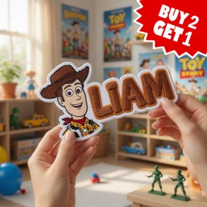 May include: A fabric patch featuring a cartoon cowboy character wearing a brown hat and a yellow shirt with a red neckerchief, next to the name LIAM in brown embroidered letters with a white border, against a white background.