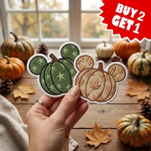 May include: Two embroidered patches shaped like pumpkins with mouse ears. One patch is dark green with light green stars, and the other is tan with brown leaf patterns. Both have white borders. A red graphic in the corner reads BUY 2 GET 1.