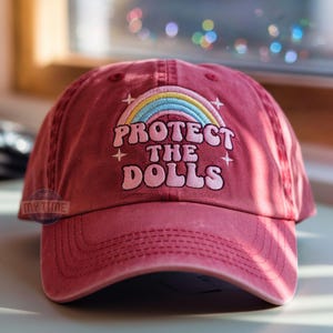 May include: A red baseball cap with a rainbow and the text "Protect the Dolls" embroidered on the front.
