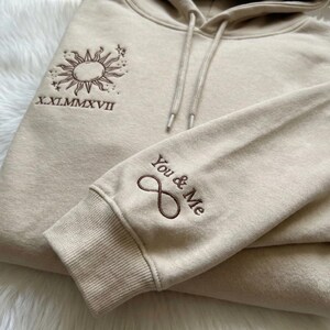 May include: Beige hoodie with a drawstring hood and ribbed cuffs. The left sleeve features the embroidered text "You & Me" with an infinity symbol. The chest has an embroidered sun and star design with the Roman numerals "X.XI.MMXVII".