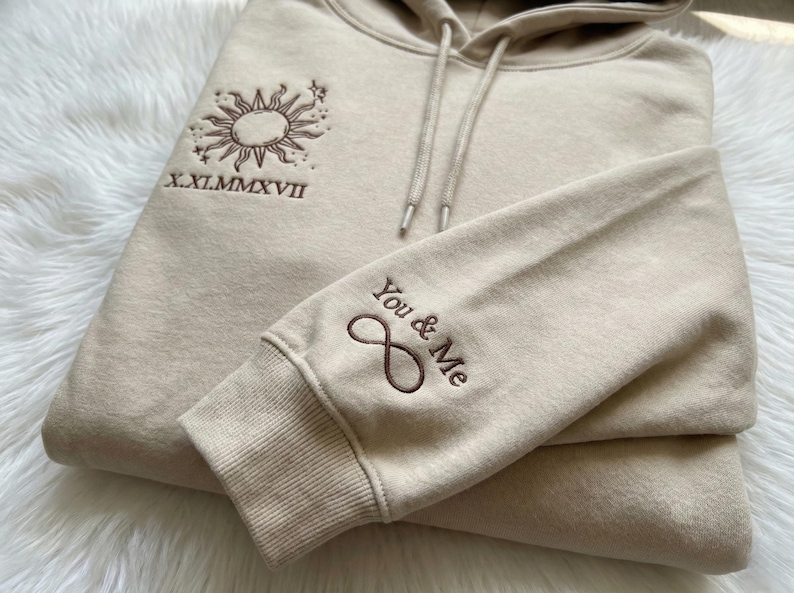 May include: Beige hoodie with a drawstring hood and ribbed cuffs. The left sleeve features the embroidered text "You & Me" with an infinity symbol. The chest has an embroidered sun and star design with the Roman numerals "X.XI.MMXVII".