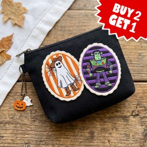 May include: A black zippered pouch featuring two oval embroidered patches: one with a ghost wearing a cowboy hat and boots against orange stripes, and another with a space ranger character against purple stripes. A small pumpkin and ghost charm hangs from the zipper.