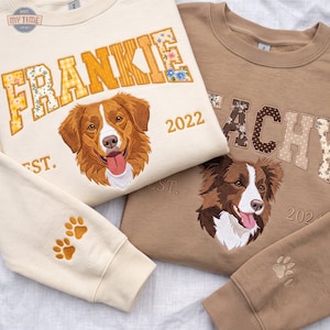 May include: Two crewneck sweatshirts in cream and tan, each featuring embroidered dog portraits, the text FRANKIE and EACHY, the years 2022 and 2024, and EST. lettering. The cream sweatshirt sleeve displays two orange paw prints, while the tan sleeve has one.