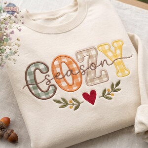 May include: A cream-coloured crewneck sweatshirt featuring embroidered text that reads COZY in large, multi-coloured gingham letters. The word season is stitched in cursive across the centre. Below, a red heart is flanked by green leaf sprigs.
