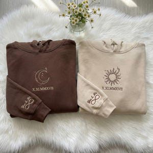 May include: Two matching hoodies, one brown and one beige, are displayed on a white fluffy surface. The brown hoodie features a crescent moon design, while the beige one has a sun design. Both have the Roman numeral date X.XI.MMXVII and the words "You & Me" with an infinity symbol.