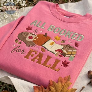 Puede incluir: Una sudadera rosa de cuello redondo con el texto bordado ALL BOOKED FOR FALL. El diseño incluye una taza de café, una calabaza, un libro abierto, una pila de libros, setas y hojas de otoño en tonos naranja, marrón y verde.