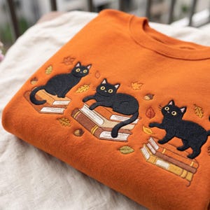 Puede incluir: Una sudadera de cuello redondo color naranja quemado con un diseño bordado de tres gatos negros sentados sobre pilas de libros. El bordado incluye pequeñas hojas de otoño amarillas y marrones y bellotas dispersas alrededor de los gatos.