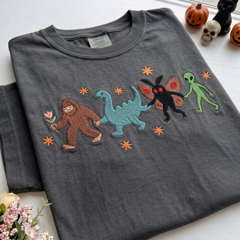 May include: A charcoal gray t-shirt featuring an embroidered design of a brown Bigfoot holding a flower, a teal dinosaur, a black moth creature with red eyes, and a green alien holding hands, surrounded by small orange stars.