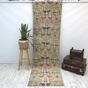 May include: A long, narrow rug with a beige background and a repeating pattern of blue, pink, and white floral motifs. The rug is hanging on a wall.