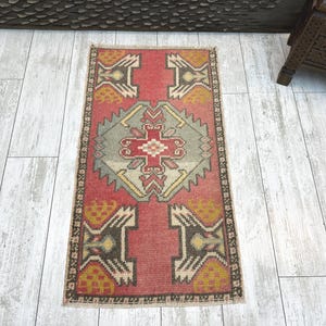 May include: A rectangular woven rug featuring a central geometric medallion in muted green, red, and yellow tones. The rug has a deep red background with symmetrical dark brown and yellow patterned motifs at the corners and edges.