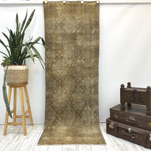 May include: A long, rectangular runner rug in a muted olive green tone, featuring a repeating symmetrical pattern of light beige floral and geometric motifs. The rug has a weathered, distressed texture and a short fringe along the bottom edge.