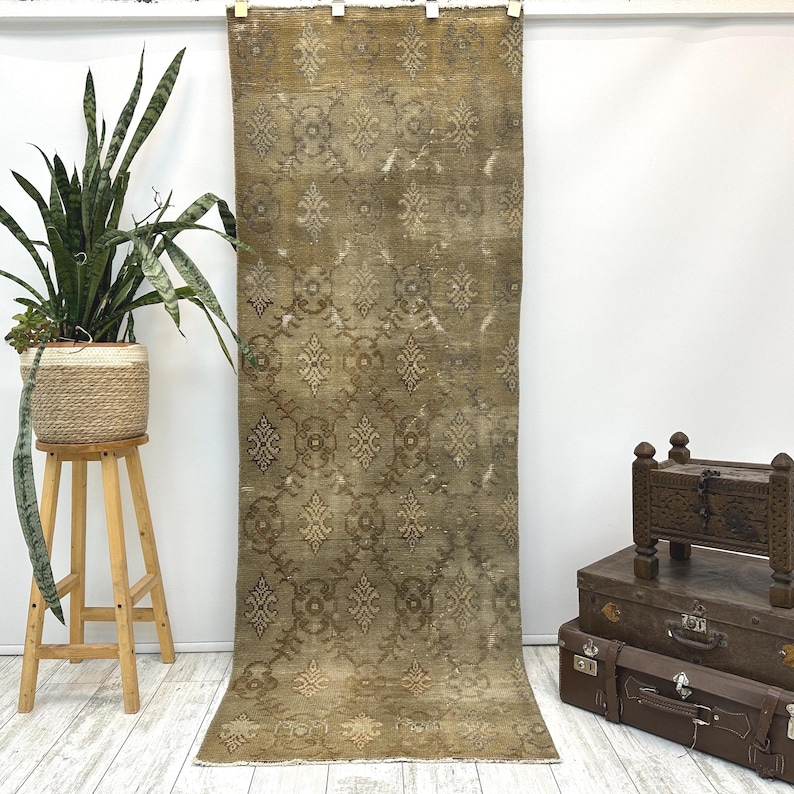 May include: A long, rectangular runner rug in a muted olive green tone, featuring a repeating symmetrical pattern of light beige floral and geometric motifs. The rug has a weathered, distressed texture and a short fringe along the bottom edge.