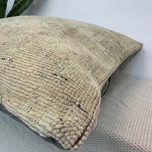May include: A square throw pillow featuring a textured, woven beige and cream front panel with a faded, intricate pattern. The pillow has a solid dark gray leather or faux leather back panel, providing a contrast in both color and material texture.