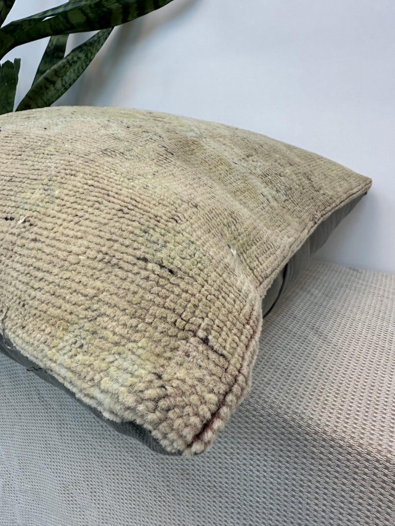 May include: A square throw pillow featuring a textured, woven beige and cream front panel with a faded, intricate pattern. The pillow has a solid dark gray leather or faux leather back panel, providing a contrast in both color and material texture.
