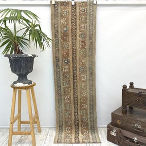 May include: A long, rectangular rug with a repeating geometric pattern in shades of brown, beige, and blue. The rug is hanging against a white wall. The rug's design features vertical stripes with square and floral motifs.