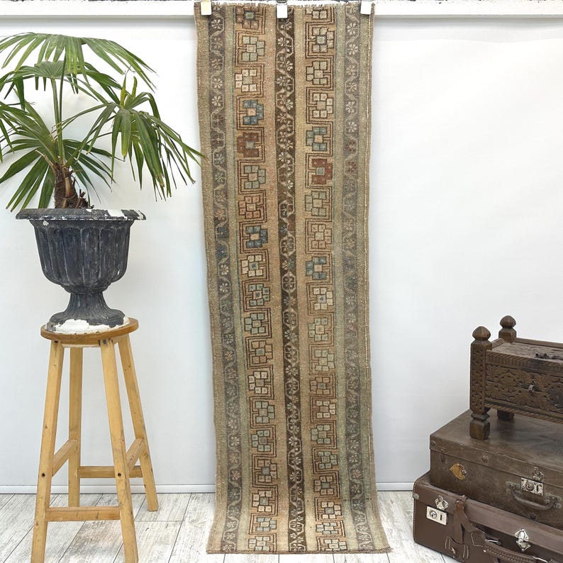 May include: A long, rectangular rug with a repeating geometric pattern in shades of brown, beige, and blue. The rug is hanging against a white wall. The rug's design features vertical stripes with square and floral motifs.