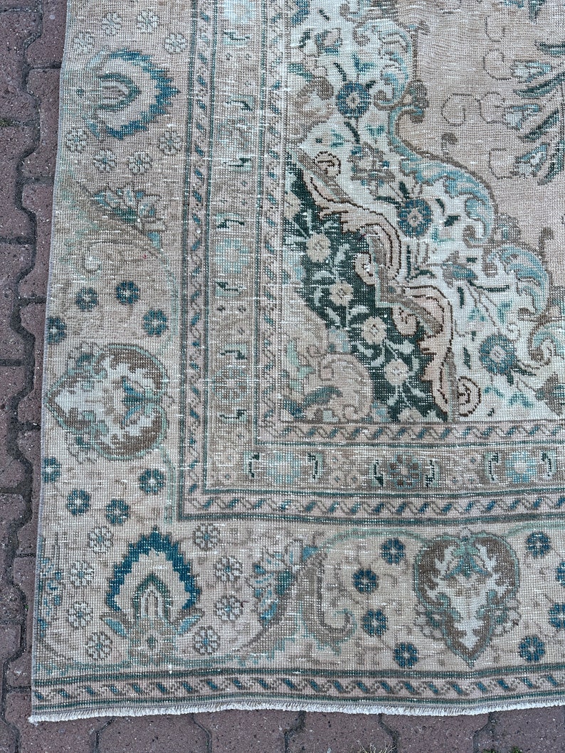 Puede incluir: Una alfombra vintage con un fondo crema descolorido y un patr&oacute;n floral azul y verde. La alfombra tiene un borde deshilachado y un aspecto desgastado.