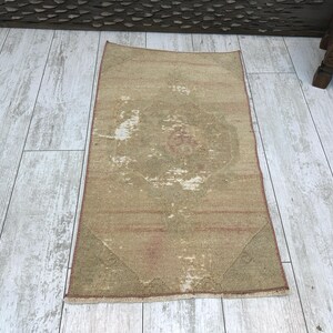 Puede incluir: Una alfombra rectangular y desgastada con una paleta de colores apagados. La alfombra presenta un diseño de medallón central en tonos beige, marrón y rojo descolorido. Los bordes y las esquinas muestran desgaste, lo que se suma a su estética vintage. La alfombra está sobre un suelo de madera clara.