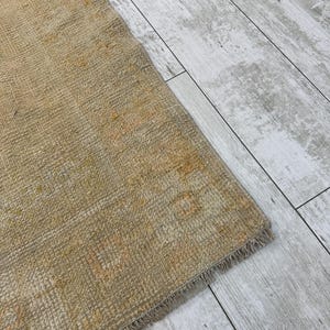 May include: Close-up of a beige rug with a fringe edge. The rug has a subtle pattern and a worn, vintage look.