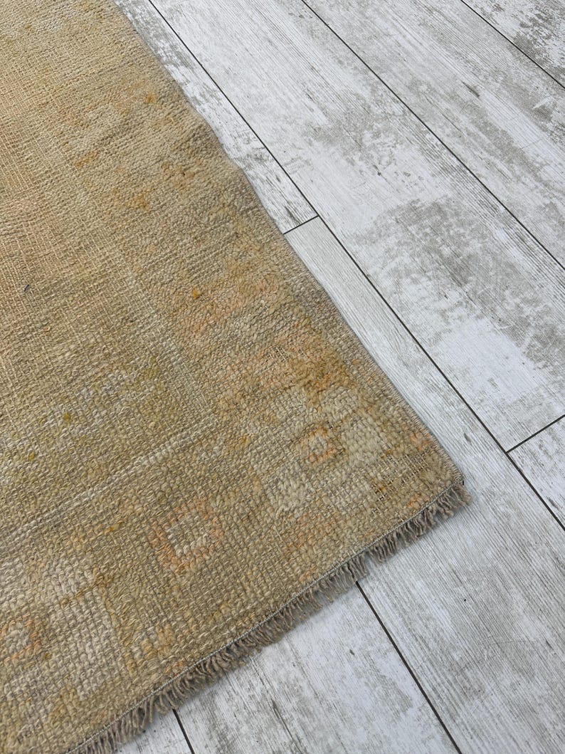 May include: Close-up of a beige rug with a fringe edge. The rug has a subtle pattern and a worn, vintage look.