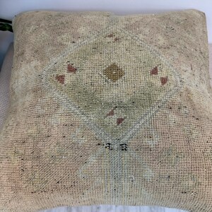 May include: A square throw pillow featuring a textured, woven surface in shades of beige, tan, and light green. The center displays a faded, geometric diamond pattern with small, muted red accents and a central brown dot, creating a worn appearance.